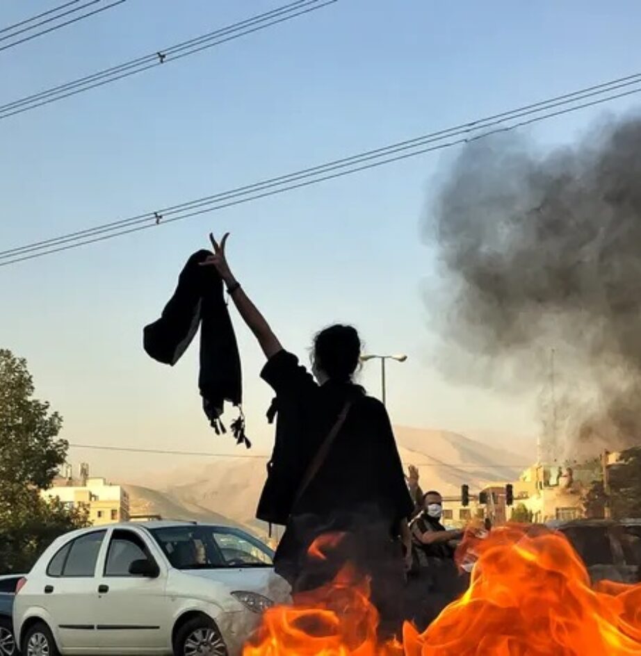 This image captures a protester raising a victory sign and holding a headscarf near a street fire during the "Woman, Life, Freedom" anti-government demonstrations in Iran, which erupted following the death of Mahsa Amini in late 2022